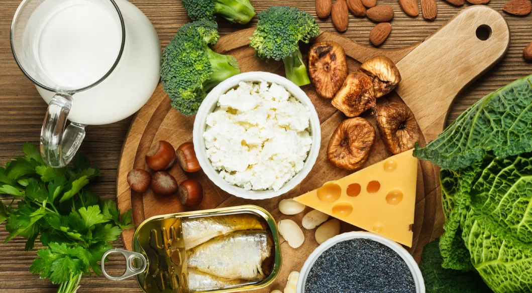 Potraviny bohaté na vápnik vrátane mlieka, syra, brokolice, sardiniek, mandlí, fíg, listovej zeleniny a tvarohu vystavené na drevenej doske.